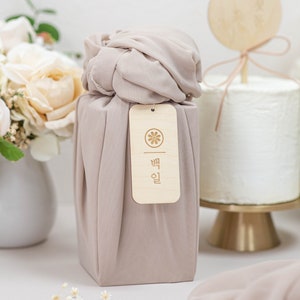 May include: A light brown gift box wrapped in a light beige fabric with a wooden tag that says "빰 인" in Korean. The box is sitting on a white surface with a small bouquet of white flowers in the foreground.
