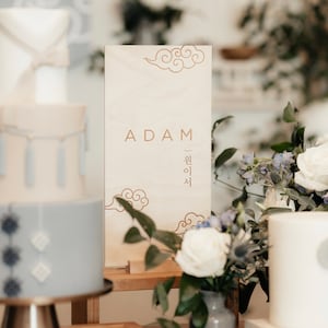 May include: A wooden sign with the name "ADAM" and Korean text in gold lettering. The sign is decorated with a carved cloud design and sits on a wooden stand. The sign is in front of a white background with a cake and flowers.