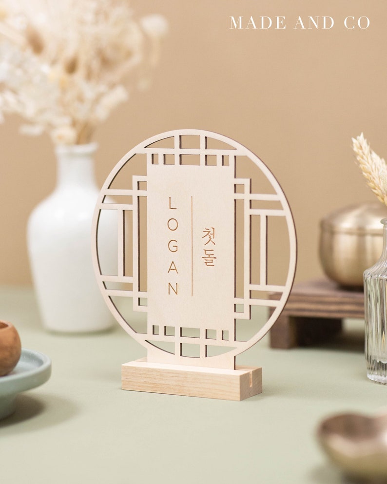 May include: A wooden table number with a circular design and a rectangular insert. The insert has the name "LOGAN" and the Korean word for "Autumn" written in gold lettering.