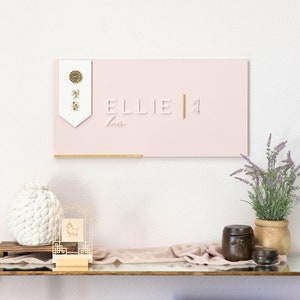 May include: A pink wooden sign with the name "Ellie" written in white letters. The sign also features a gold Chinese character and a gold symbol. The sign is hanging on a white wall above a shelf with decorative items.