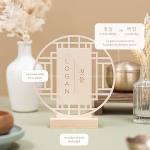 May include: A wooden sign with a traditional Korean design, featuring a customizable first name and the text "첫돌" (meaning "first birthday") or "백일" (meaning "100th day"). The sign comes with a wooden stand.