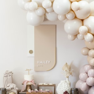 May include: A first birthday party table setting with a white and beige balloon arch, a cake with a gold "one" topper, and a sign that says "HAILEY" in white lettering with the Korean word for "one" underneath. The table is decorated with a variety of items, including a white ceramic pineapple, a gold bowl, and a wicker basket.