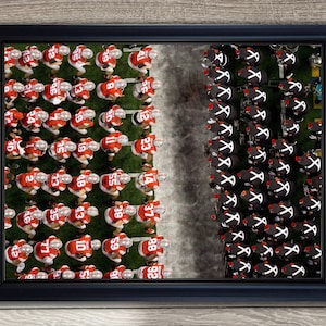 May include: Framed aerial view of a football game. Two teams face each other on a green field, one in red and white uniforms, the other in black and white. The image is framed in a black border.