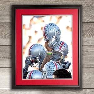 May include: A group of football players wearing gray helmets with white stars. The helmets are stacked on top of each other. The players are wearing black jerseys and white pants. The background is a blurred image of a crowd of people.