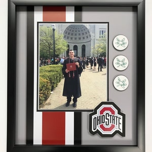 May include: A black framed photo of a person in a graduation gown holding a diploma in front of a building. The photo is surrounded by a gray mat with three white circles with green leaf designs and a black and red Ohio State logo.