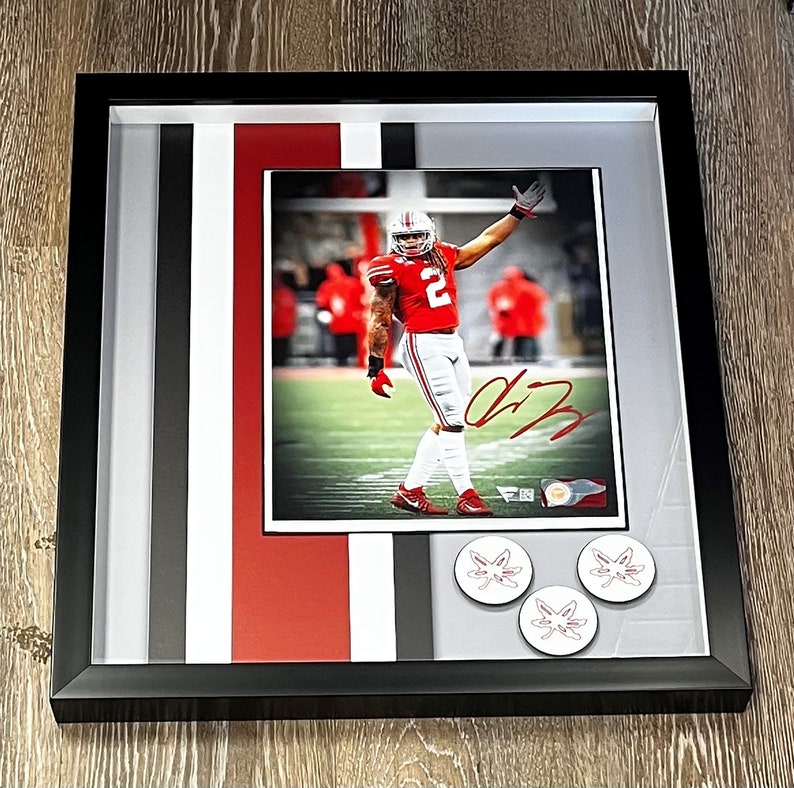 K&ouml;nnte beinhalten: Ein gerahmtes Foto eines Footballspielers, der ein rot-wei&szlig;es Trikot mit der Nummer 2 tr&auml;gt. Der Spieler signiert seinen Namen auf dem Foto. Der Rahmen hat einen schwarzen Rand und einen wei&szlig;en Passepartout mit roten, schwarzen und wei&szlig;en Streifen. In der rechten unteren Ecke des Rahmens befinden sich drei wei&szlig;e runde Aufkleber mit einem roten Ahornblatt-Design.