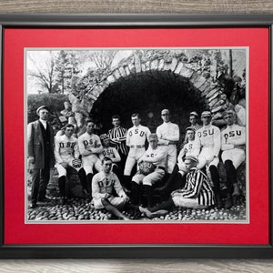 May include: A black and white photograph of a group of men in athletic uniforms. The men are standing and sitting in front of a stone archway. The men are wearing jerseys with the letters "OSU" on them. The photograph is framed in a black frame with a red mat.