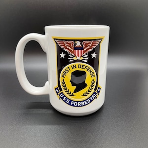 May include: White ceramic mug with a black and gold design featuring an eagle, arrows, stars, and the text "First in Defense U.S.S. Forrestal".