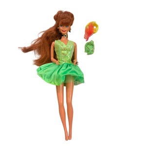 Puede incluir: Una muñeca con largo cabello castaño rojizo viste un chaleco verde lima sin mangas con tres botones y una falda acampanada a juego. La muñeca lleva una gargantilla negra con un colgante dorado y pendientes dorados. Incluye un accesorio amarillo y naranja.