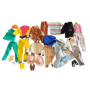Op de afbeelding: Een verzameling poppenkleding en accessoires, waaronder een groene outfit, een neongele en roze jas, een bruine jas met franjes, een bruine broek, een gestreept shirt, een zwarte jas, een blauwe hoed, een geruite broek, een lichtblauwe badjas en twee cowboyhoeden.