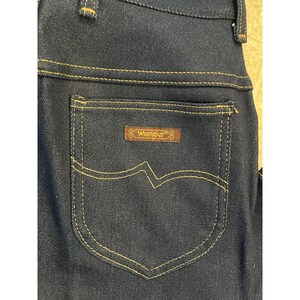 May include: Close-up of dark blue denim jeans with a back pocket. The pocket has a decorative stitch design and a brown rectangular patch with the brand name "Wrangler" in gold lettering.