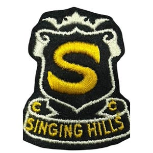 May include: A black and gold embroidered patch with the words "SINGING HILLS" at the bottom. The patch features a shield design with a large yellow "S" in the center. The border is white.