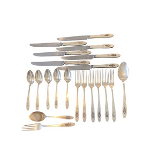 May include: A collection of vintage silverware, including knives, forks, and spoons, arranged on a white surface. The knives have cream-colored handles and ornate detailing. The forks and spoons are silver-toned with decorative patterns.