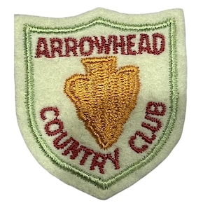 Puede incluir: Un parche de fieltro en forma de escudo con una base verde claro y un borde bordado en verde claro. El centro presenta un icono de punta de flecha bordado en oro. El texto ARROWHEAD está bordado en rojo arriba, COUNTRY CLUB en rojo abajo.