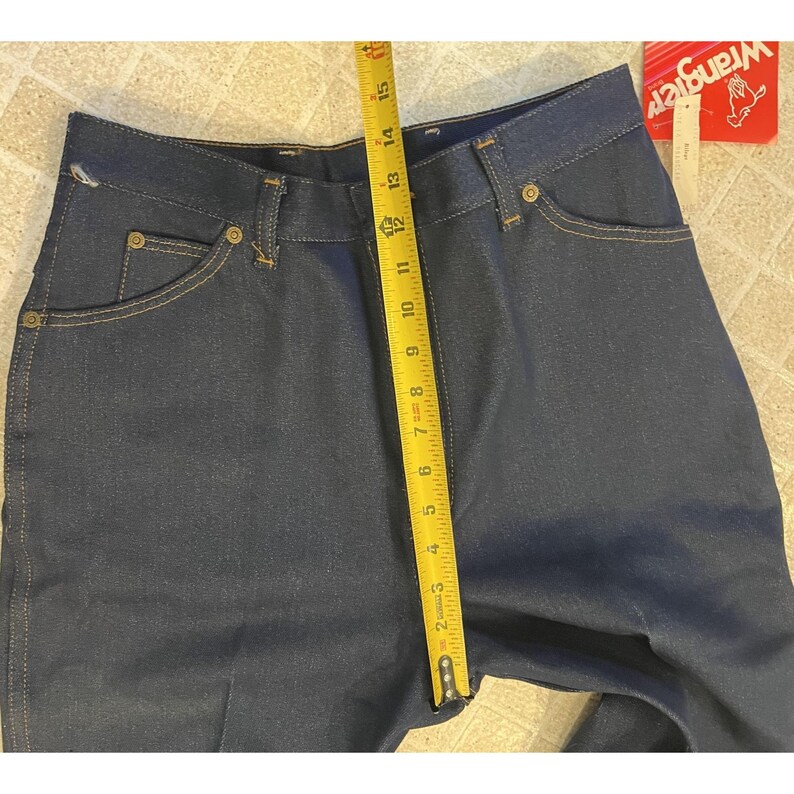 May include: Dark blue denim jeans with a yellow measuring tape showing a length of approximately 15 inches. The jeans have classic five-pocket styling and a Wrangler tag.