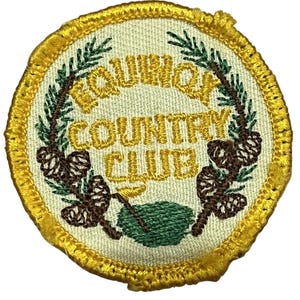 May include: Circular embroidered patch featuring the text "EQUINOX COUNTRY CLUB" in gold thread. The patch has a cream-coloured centre and a gold border. Pine cones and green foliage are embroidered around the text.