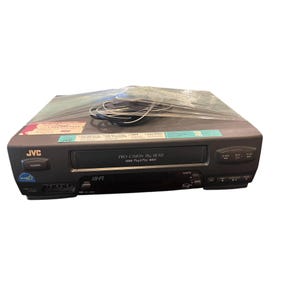 May include: A dark gray JVC VCR player featuring a Pro-Cision 19u Head system, a power button, and playback controls. The device is wrapped in clear plastic film with a coiled black power cord on top and includes various printed labels.