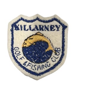 May include: An embroidered patch in white and blue, featuring the text "KILLARNEY GOLF & FISHING CLUB". The design includes a gold fish leaping from blue water. The patch is shield-shaped.