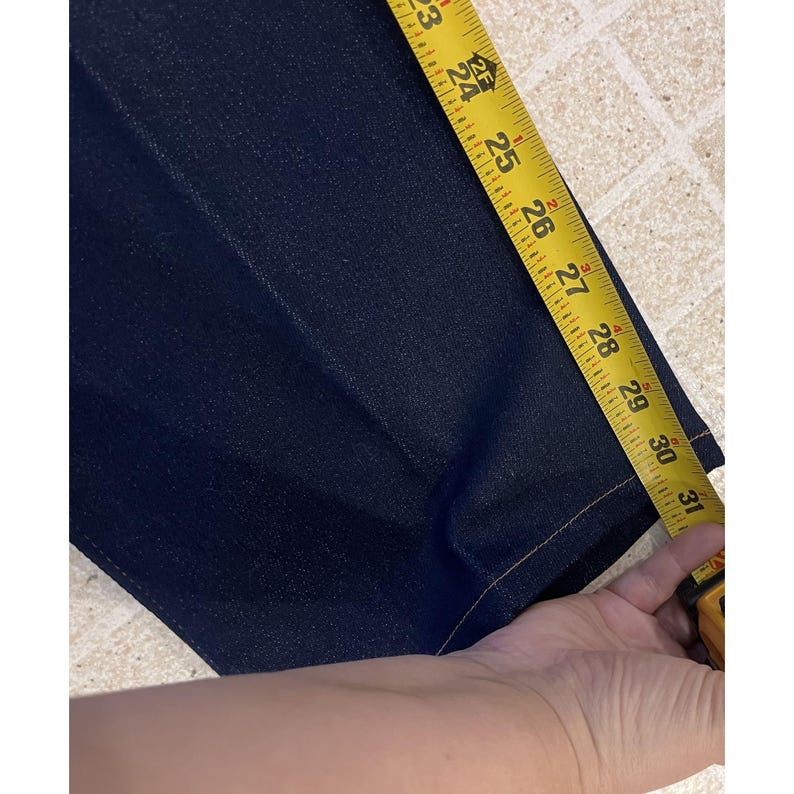 May include: Dark blue denim pants being measured with a yellow tape measure. The tape measure shows a length of approximately 23 to 31 inches. The pants have a classic five-pocket design.