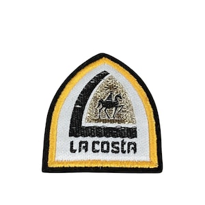 Könnte beinhalten: Ein weißer und goldener gestickter Aufnäher mit einem schwarzen Pferd- und Reitermotiv. Der Aufnäher ist bogenförmig und trägt den Text "LA COSTA" in schwarzen Buchstaben.