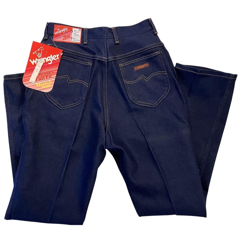May include: Dark blue Wrangler jeans with a high waist and a classic fit. The jeans have a straight leg, a Wrangler label, and a tag that reads "Misses Full Fit". The back pockets have a distinctive curved design.