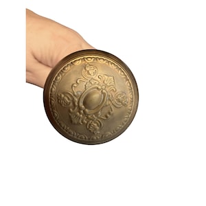 May include: A brass-colored, round doorknob with an ornate, embossed design. The central oval is surrounded by floral and scrollwork details. The doorknob is held in a hand against a white background.