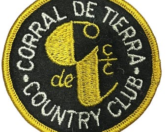 May include: Circular embroidered patch with the text "CORRAL DE TIERRA COUNTRY CLUB" in white, curving around a black circle. The centre features a gold-coloured stylized "9" and "c/t". The patch has a gold border.
