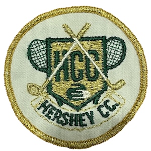 May include: Circular embroidered patch with a cream-coloured background and a gold border. The patch features a green and gold shield design with the letters "HGC" and the words "HERSHEY CC" in green. Crossed hockey sticks and tennis rackets are also included.