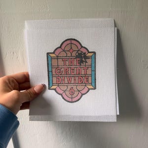 May include: A white needlepoint canvas featuring a central design with the text THE GREAT DIVIDE in pink letters. The design is framed by a pink and blue geometric border with a black outline, resembling a stained glass window pattern.