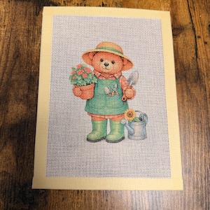 May include: A needlepoint canvas featuring a brown teddy bear dressed in a green apron, a straw-colored hat, and green boots. The bear holds a potted plant with pink flowers, while a small blue watering can with a sunflower sits nearby.