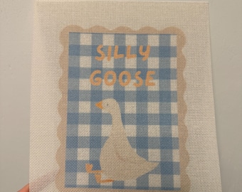 May include: A needlepoint canvas featuring a white goose with an orange beak and feet, set against a blue and white gingham background. The design is framed by a scalloped tan border and includes the text SILLY GOOSE in orange block letters.