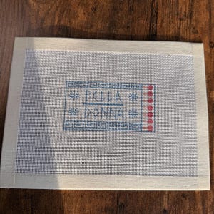 May include: A rectangular needlepoint canvas featuring a white mesh background with a cream-colored fabric border. The center displays a blue stitched design with the text BELLA DONNA, accented by small red dots and a geometric Greek key border pattern.