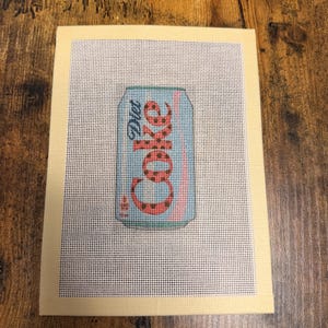 May include: A needlepoint canvas featuring a light blue soda can with the text Diet Coke in red letters with black polka dots. The design is centered on a white grid mesh background with a solid cream-colored border framing the entire rectangular piece.