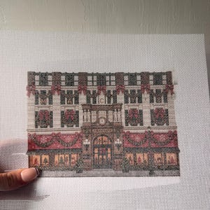 May include: A needlepoint canvas featuring a detailed architectural design of a multi-story building facade. The pattern includes rows of windows, decorative red swags, and a central arched entrance, all rendered in shades of brown, red, and black.