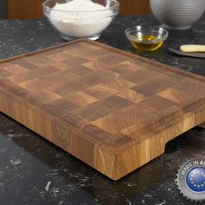 Puede incluir: Una tabla de cortar rectangular de madera con un patrón de tablero de ajedrez. La tabla tiene una ranura para jugos y un recorte para el mango. La tabla está hecha de madera marrón clara y oscura. Una pegatina "Made in Europe" es visible.