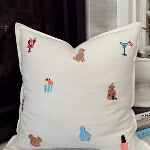 May include: A square off-white throw pillow featuring scattered embroidered icons including a red lobster, a brown dog with a blue collar, a martini glass with an American flag, a striped ice cream cup, a brown bear, a mouse head, a blue foam finger, and pink nail polish.