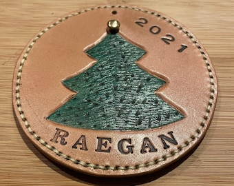 May include: A tan leather Christmas ornament with a green felt Christmas tree and the name "Raegan" and the year "2021" embossed on it.