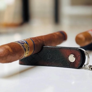 May include: A brown cigar rests on a black and red leather key holder with a silver key ring. The cigar has a blue and gold band. Another cigar is visible in the background.