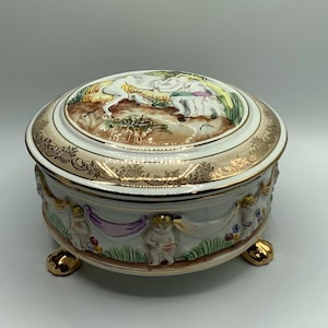 May include: A decorative porcelain trinket box with a lid. The box is white with gold accents and features a painted scene on the lid. The sides are adorned with cherubs and floral details. The box rests on three gold feet.