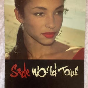 Könnte beinhalten: Werbeplakat für die "Sade World Tour". Das Bild zeigt eine Frau mit dunklem Haar und rotem Lippenstift. Der Text "Sade World Tour" ist rot und weiß auf schwarzem Grund. Im Hintergrund eine Strandszene.