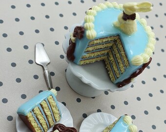 May include: A miniature blue and white cake with chocolate accents and a bunny on top, displayed on a white cake stand with two smaller cakes on white plates and two silver forks.