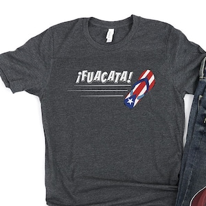 May include: A dark gray t-shirt with the text "&iexcl;FUACATA!" and a red, white, and blue flip-flop with a star on it. The t-shirt is paired with blue jeans and maroon high-top sneakers.