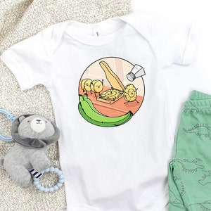 May include: White baby onesie with a cartoon illustration of plantains being sliced with a knife and sprinkled with salt. The illustration is in a circle with a green plantain on the bottom.