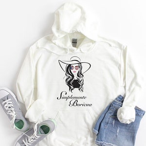 May include: Cream-colored hoodie with a graphic of a woman in a hat and sunglasses, with the text "Simplemente Boricua." The hoodie is paired with denim shorts and gray sneakers. The image is set against a white background.