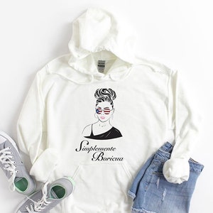 May include: White hoodie with a black and white illustration of a woman with a bun in her hair wearing sunglasses with the American flag on them. The text "Simplemente Boricua" is printed below the illustration.