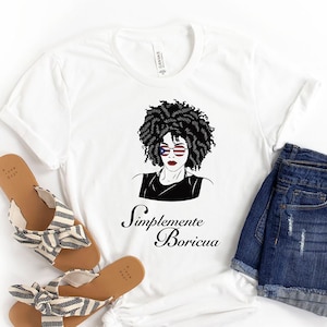 May include: White t-shirt with a black and white illustration of a woman with curly hair wearing sunglasses with the American flag design. The text "Simplemente Boricua" is printed below the illustration.