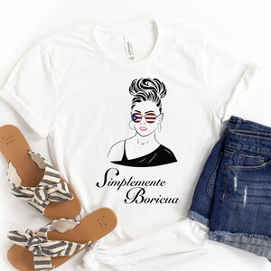 May include: White t-shirt with a black and white illustration of a woman with a bun wearing sunglasses with the American flag design. The text "Simplemente Boricua" is printed below the illustration.