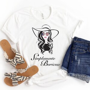 May include: White t-shirt with a black and white illustration of a woman wearing a hat and sunglasses with an American flag design. The text "Simplemente Boricua" is printed below the illustration.