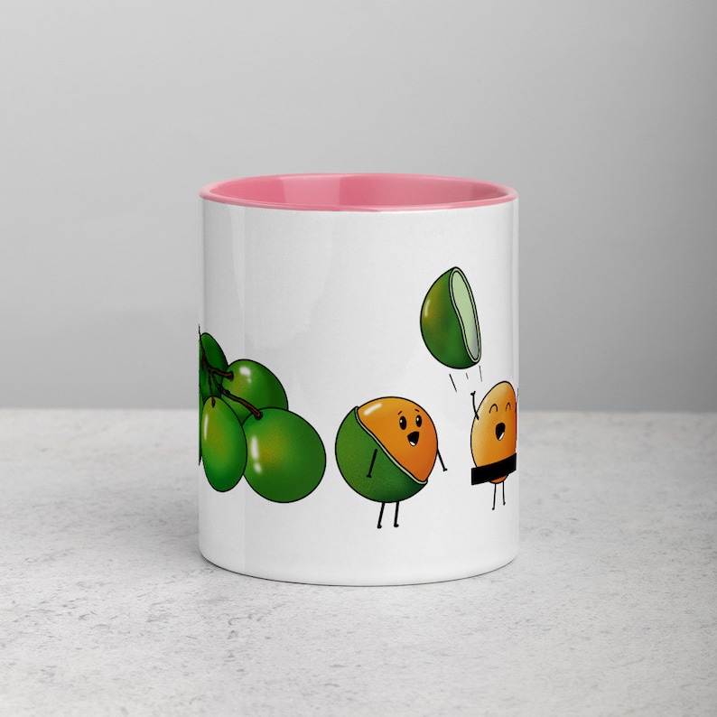 May include: White ceramic mug with a pink rim. The mug features a cartoon illustration of green limes and a lime with a face throwing another lime with a face. The lime with a face is wearing a brown belt.