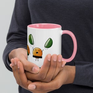 May include: White ceramic mug with a pink rim and handle. The mug features a cartoon illustration of a smiling orange with two green halves of a lime on either side of it. The orange is holding the lime halves up in the air.